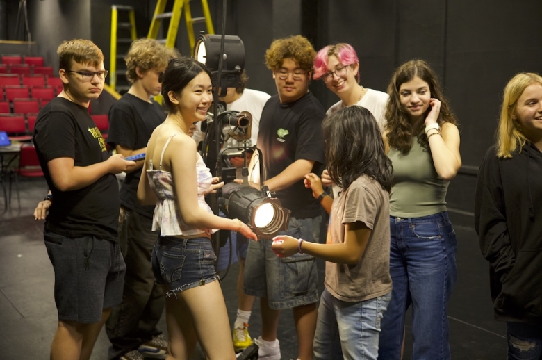 Group of students stand, smiling, around a spotlight during a Drama: Production & Design class